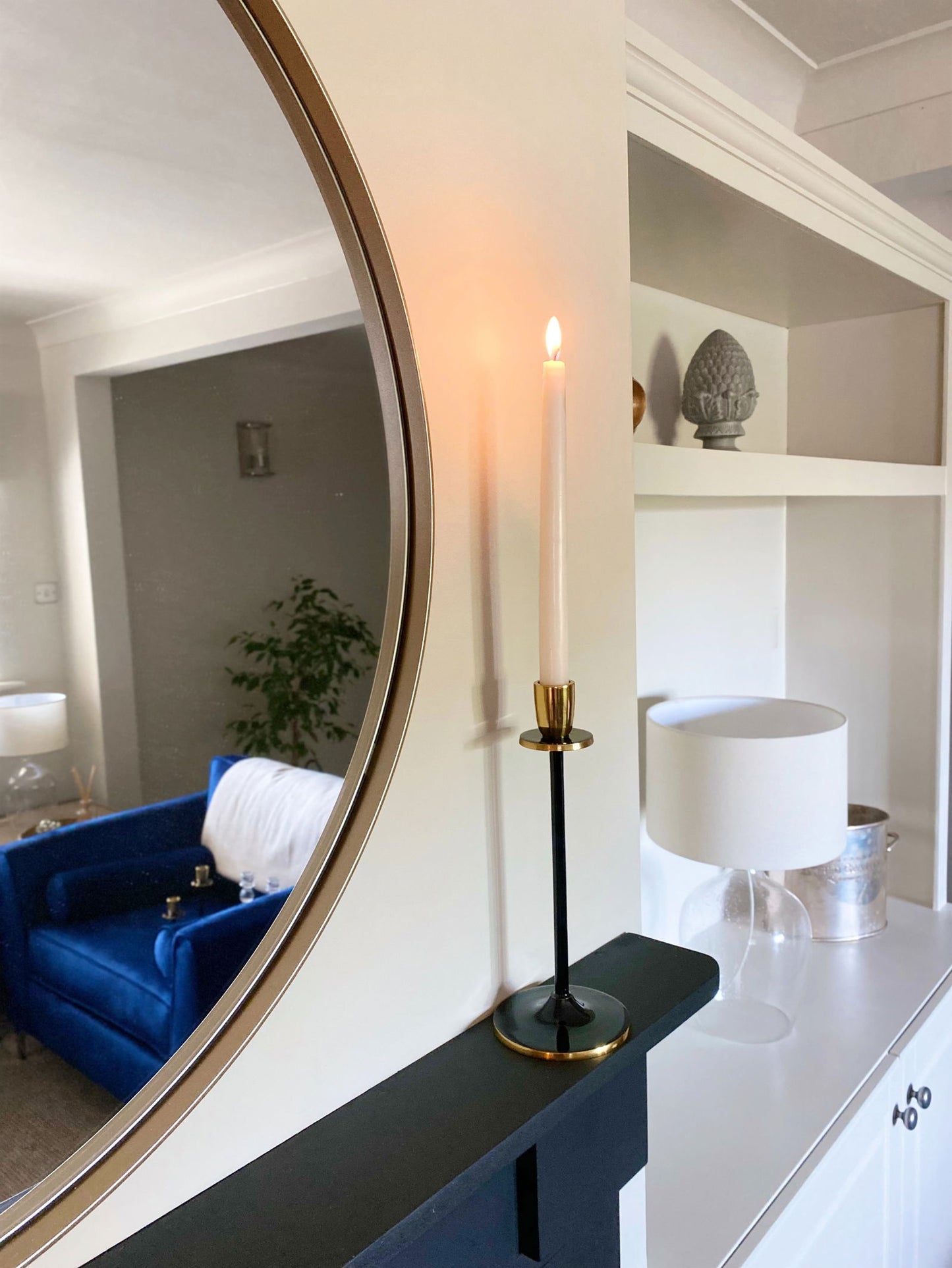 Black and gold candlestick holding a lit white taper candle on a black shelf near a round mirror
