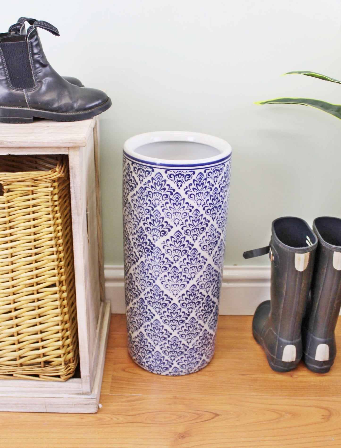 Blue and white umbrella holder with intricate pattern placed on wooden floor between boots and wicker basket