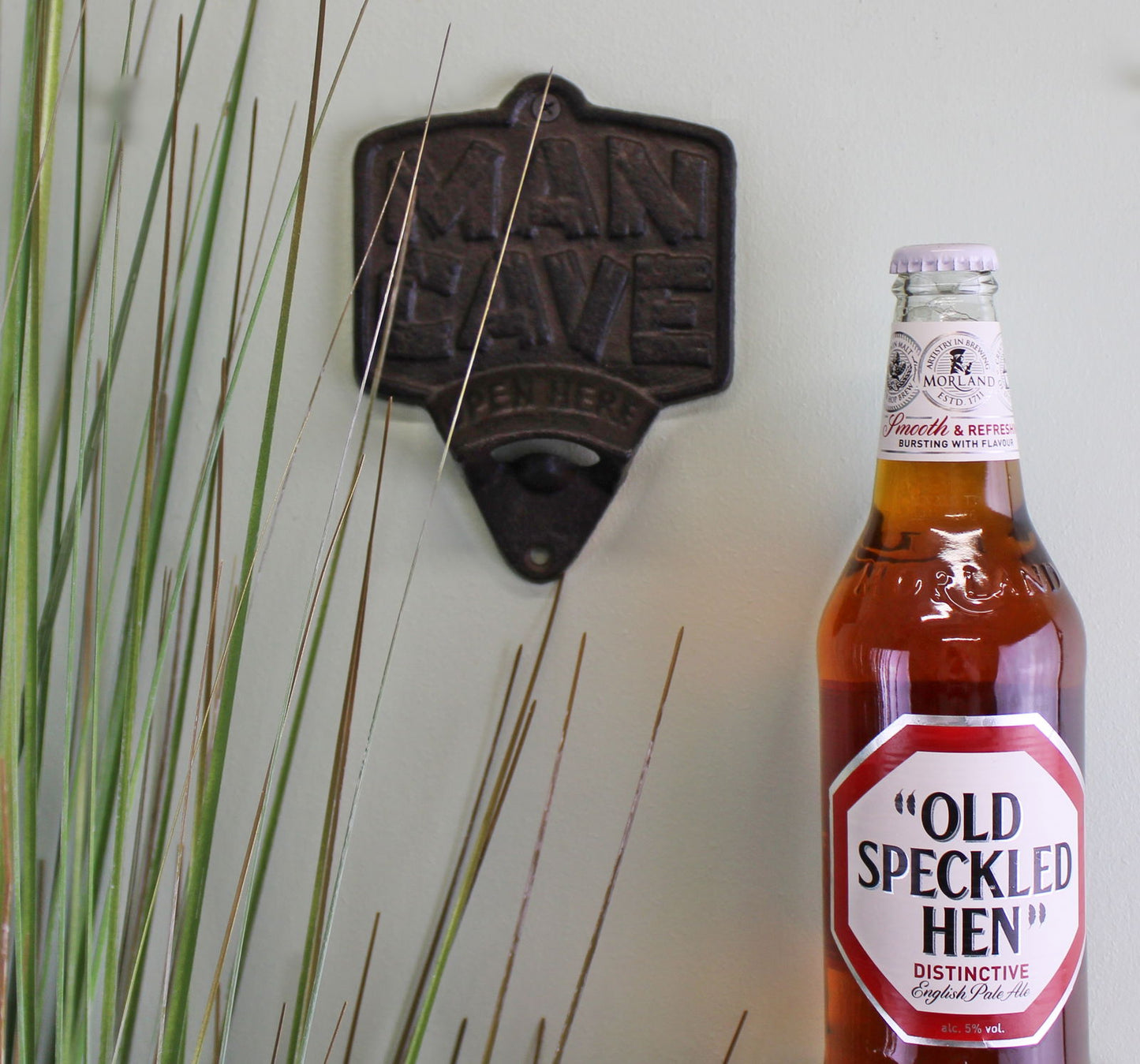 Geko Products cast iron bottle opener wall mount with rustic design on white wall next to a brown Old Speckled Hen beer bottle
