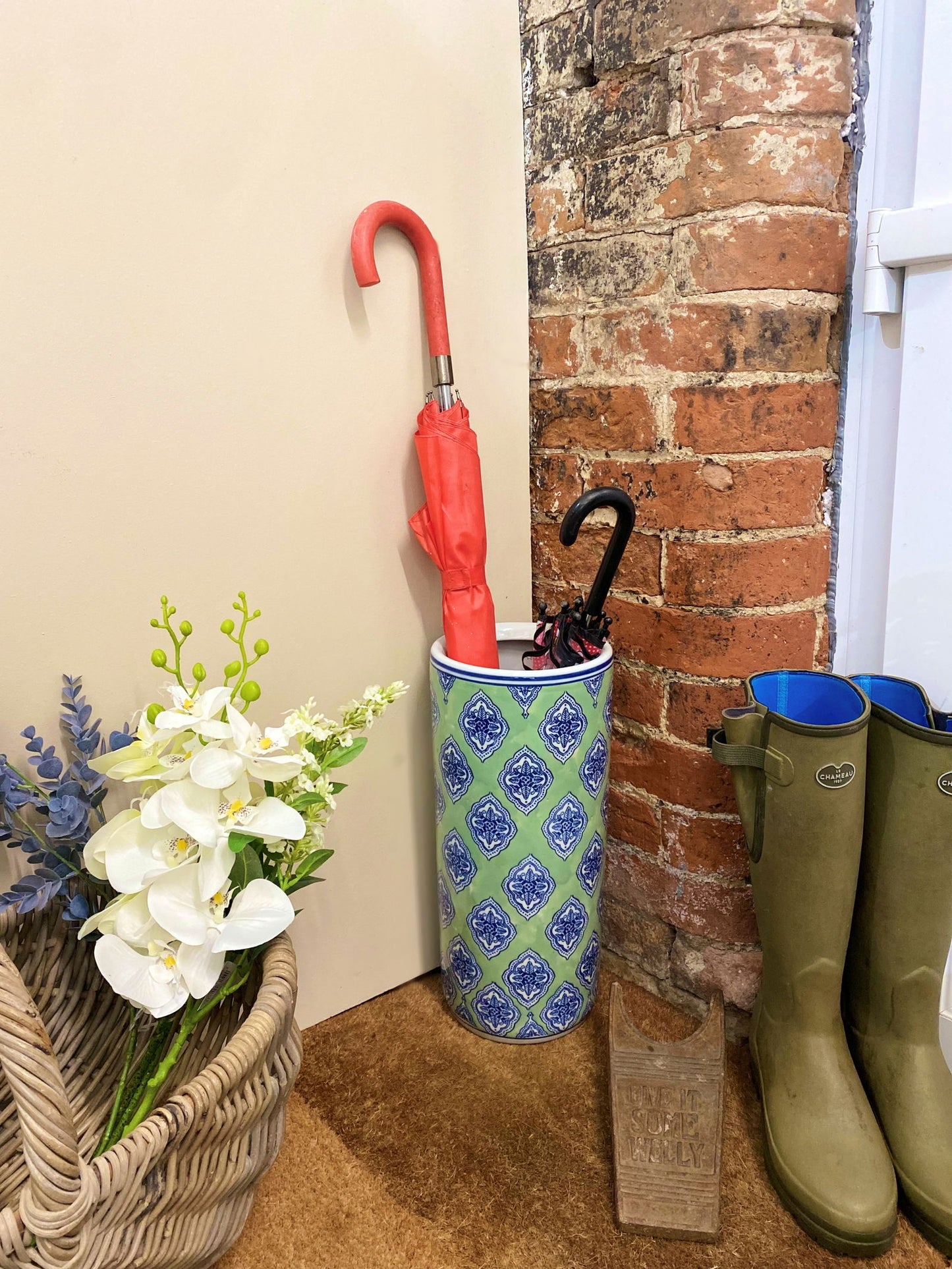 Geko Products green diamond umbrella stand holding umbrellas by a brick wall next to green boots and a basket of flowers