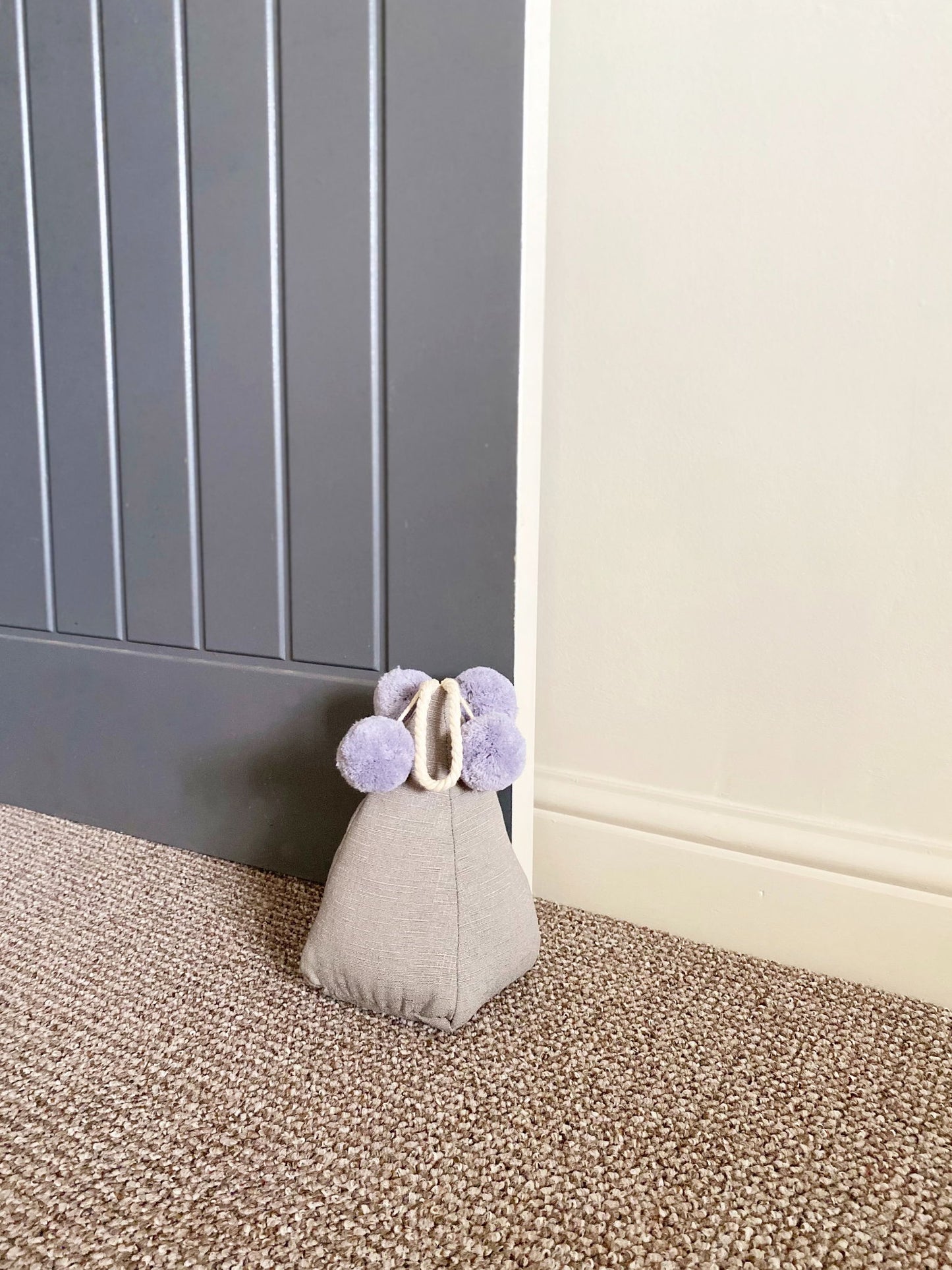 Grey pompom doorstop holding a gray door open on carpeted floor by white wall