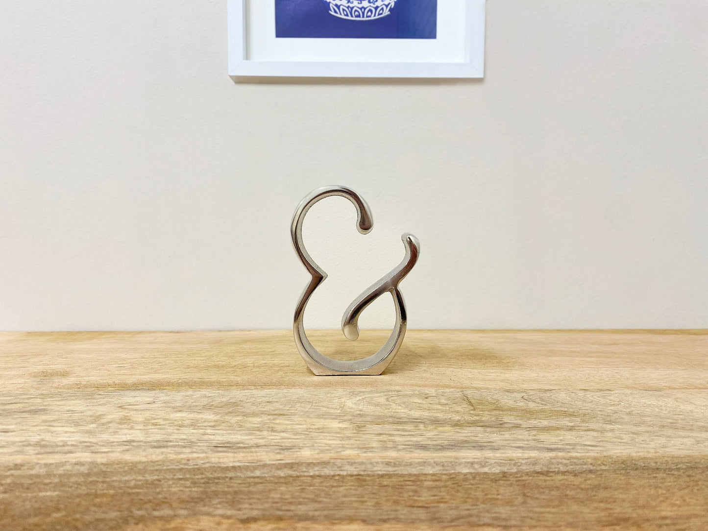 Silver ampersand home decor piece on wooden surface with framed artwork in background