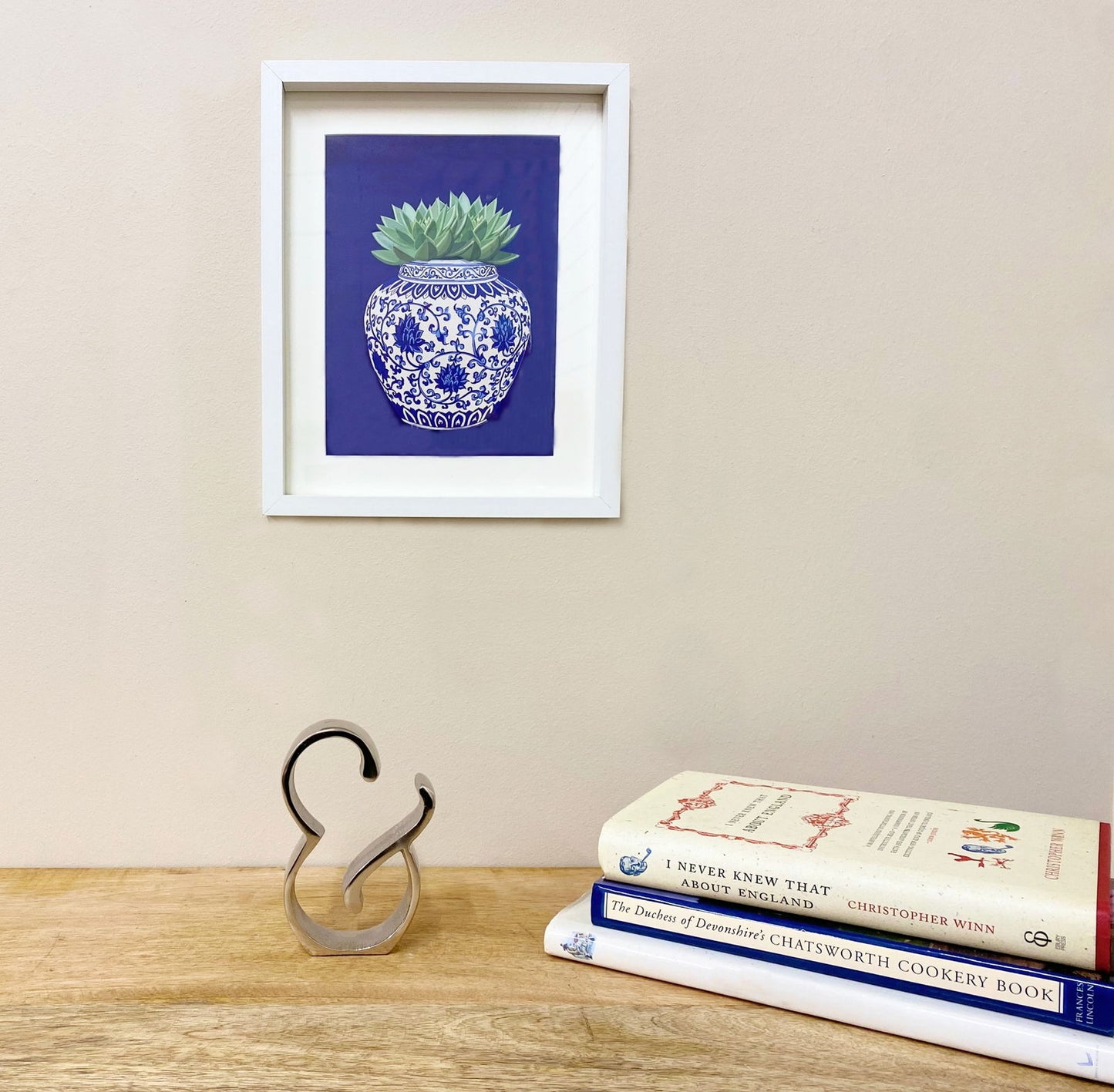 Silver ampersand home decor sculpture displayed on wooden surface next to stacked books and framed plant artwork
