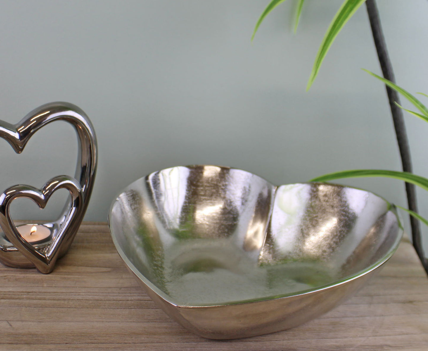 Silver heart decorative bowl with a shiny finish placed on a wooden surface next to a heart-shaped candle holder
