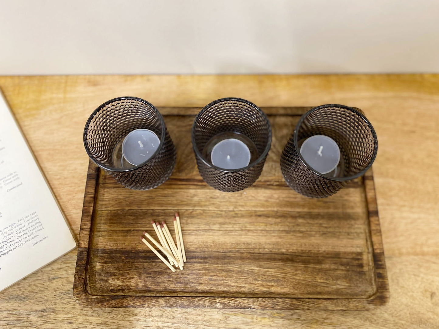 Three black mesh spiritual tea light holders with white candles on a wooden tray next to matches