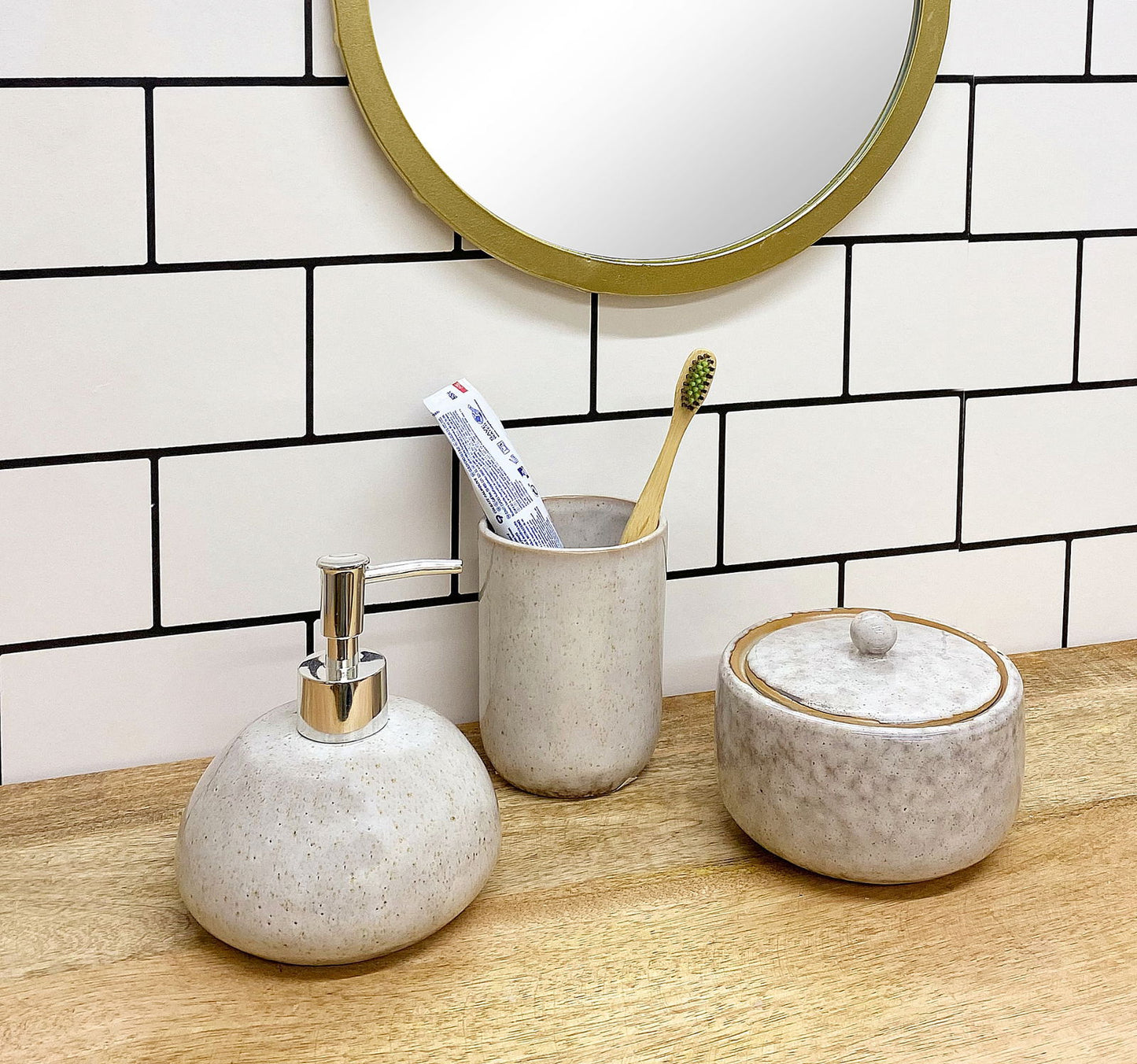 Taupe ceramic soap dispenser with silver pump next to matching toothbrush holder and lidded jar on wooden counter
