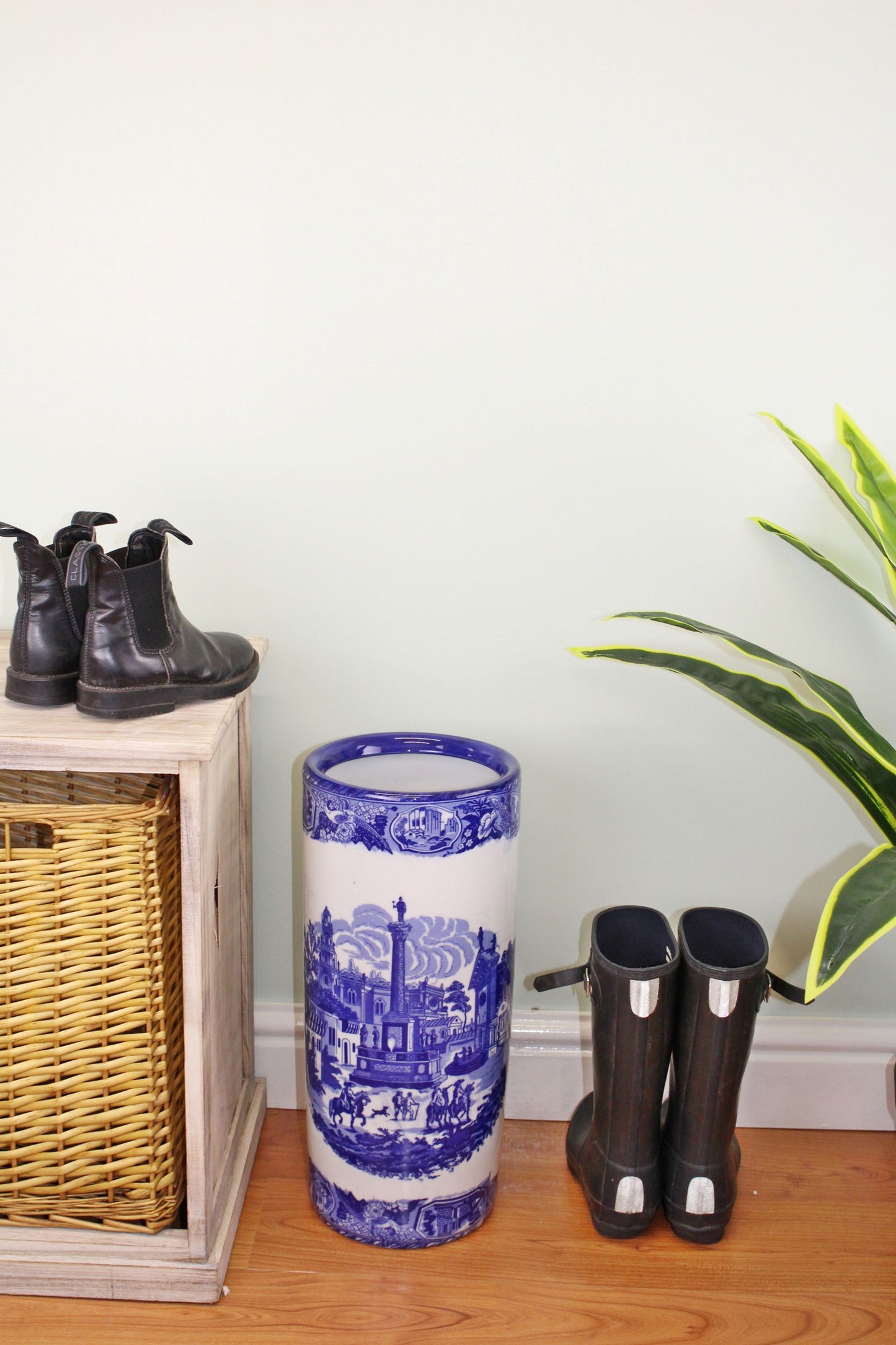 Geko Products blue and white ceramic townscape design umbrella holder placed beside black boots and a wicker cabinet
