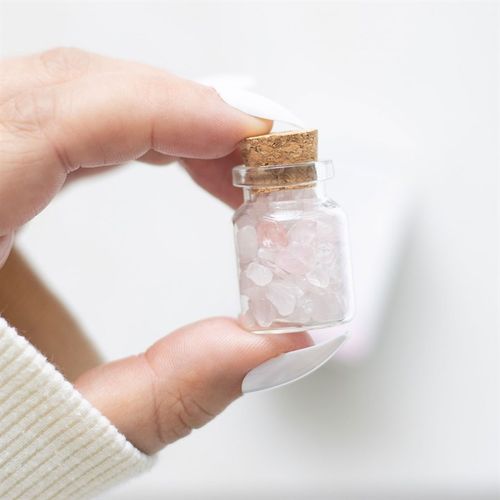 Small jar of rose quartz in matchbox held between fingers, showcasing clear glass and cork top with pink crystal pieces