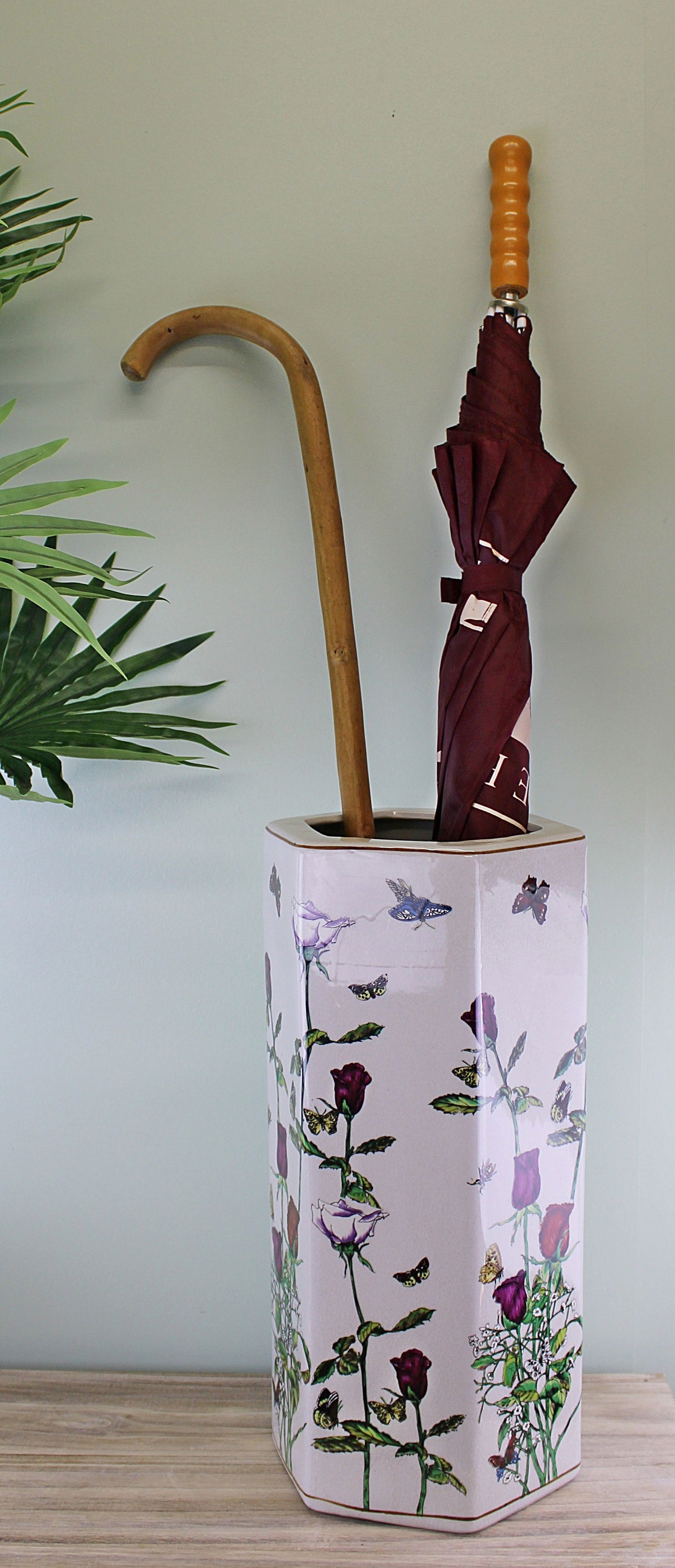 Hexagonal ceramic umbrella stand with floral and butterfly design holding a cane and a folded maroon umbrella