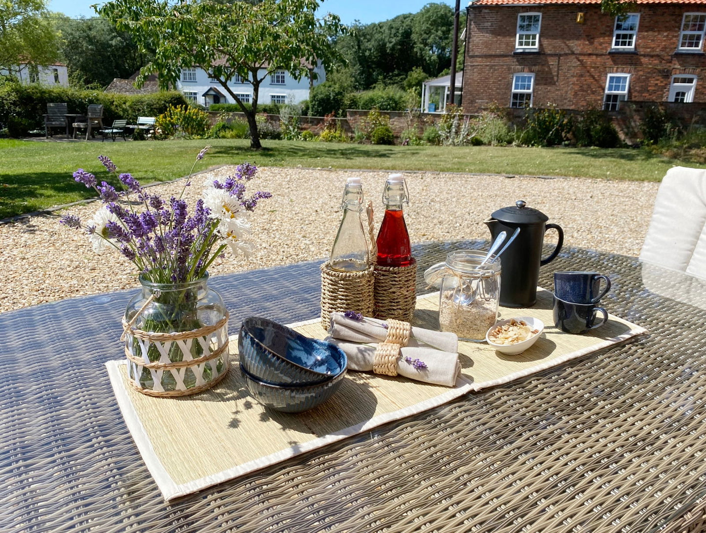 Outdoor dining table with woven grass place mats set, lavender bouquet, glass bottles, and tableware on sunny patio