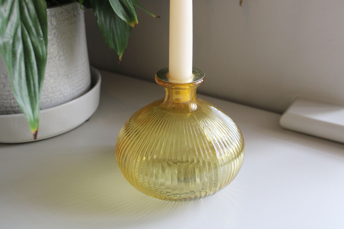 Yellow ribbed glass candle holder by Geko Products with a white candle on a white surface near a plant