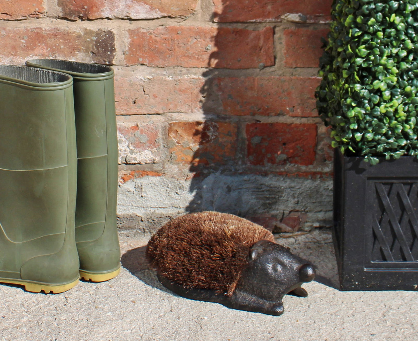 Cast iron garden decor hedgehog doorstop with brown bristles placed outside near green boots and plant pot
