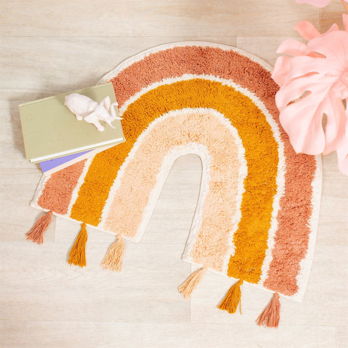 Earth rainbow rug with tassels in warm earthy tones placed on wooden floor with books and pink plant