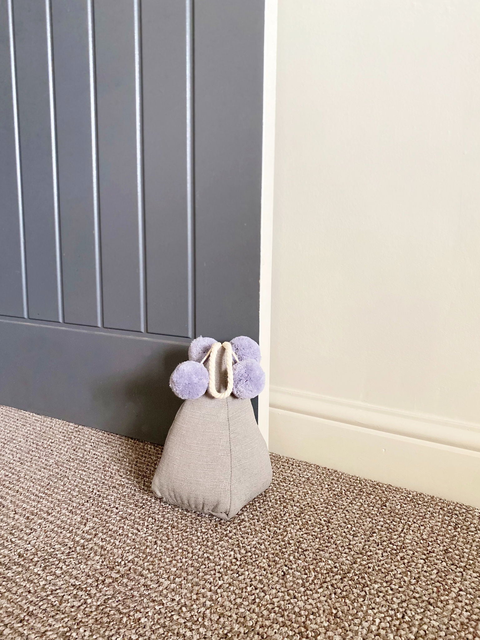 Grey pompom doorstop holding a gray door open on carpeted floor by white wall