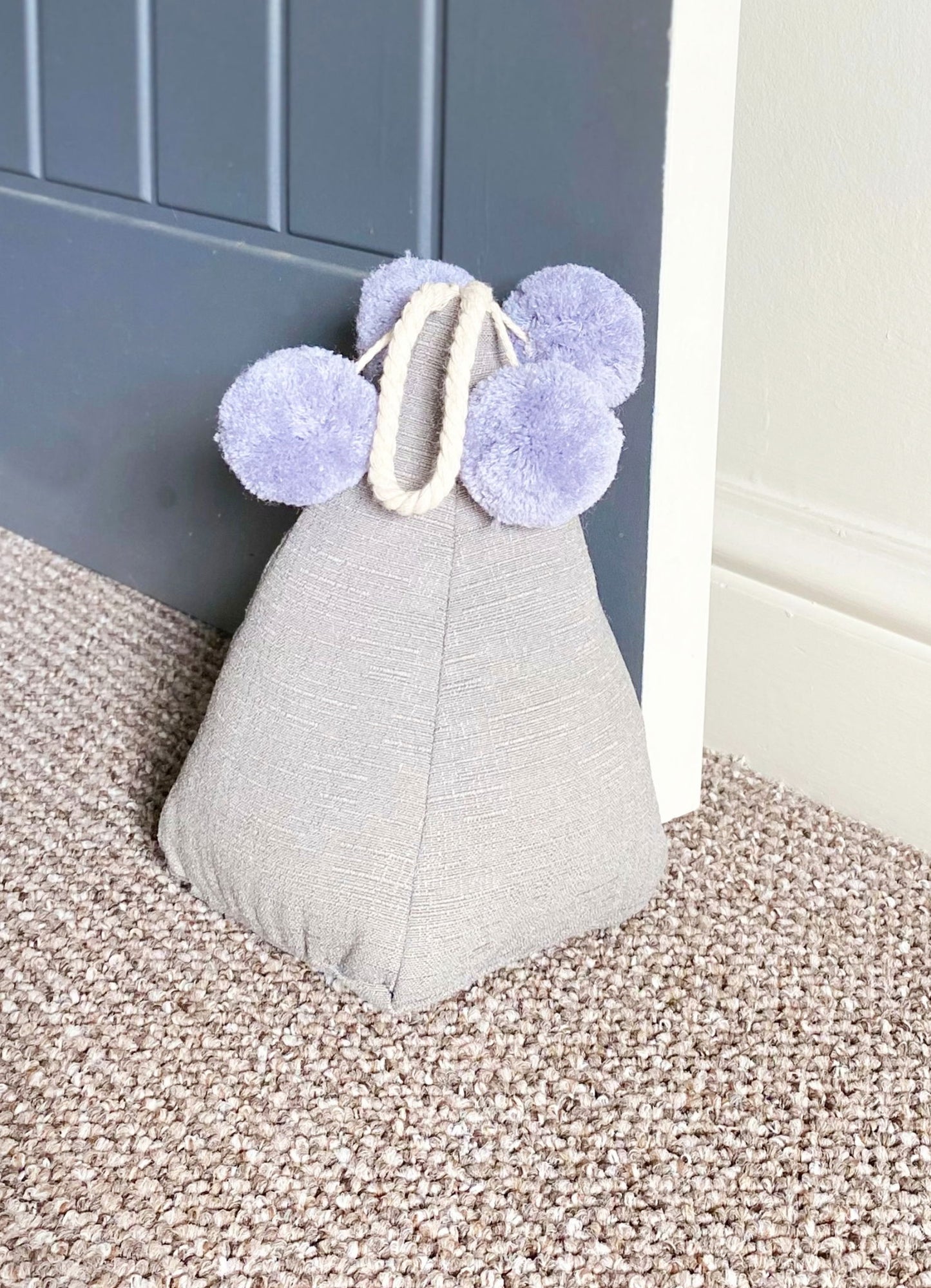 Geko Products grey pompom doorstop with fabric texture placed on carpet by a blue door